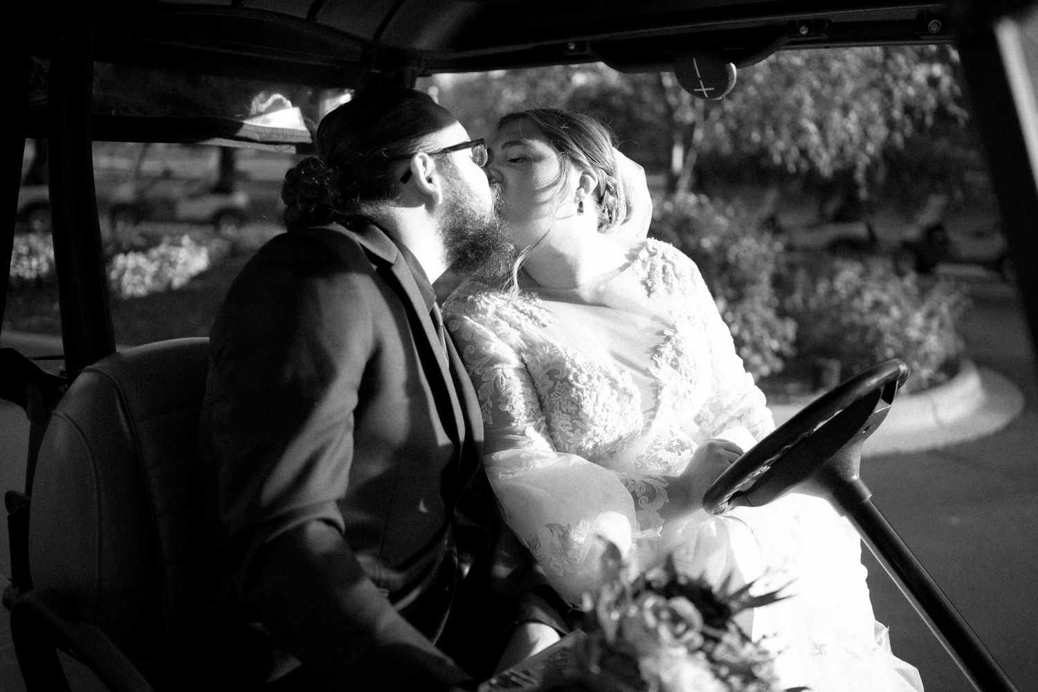 A romantic black and white photo of newlyweds sharing a kiss in the driver's seat of their vintage car.