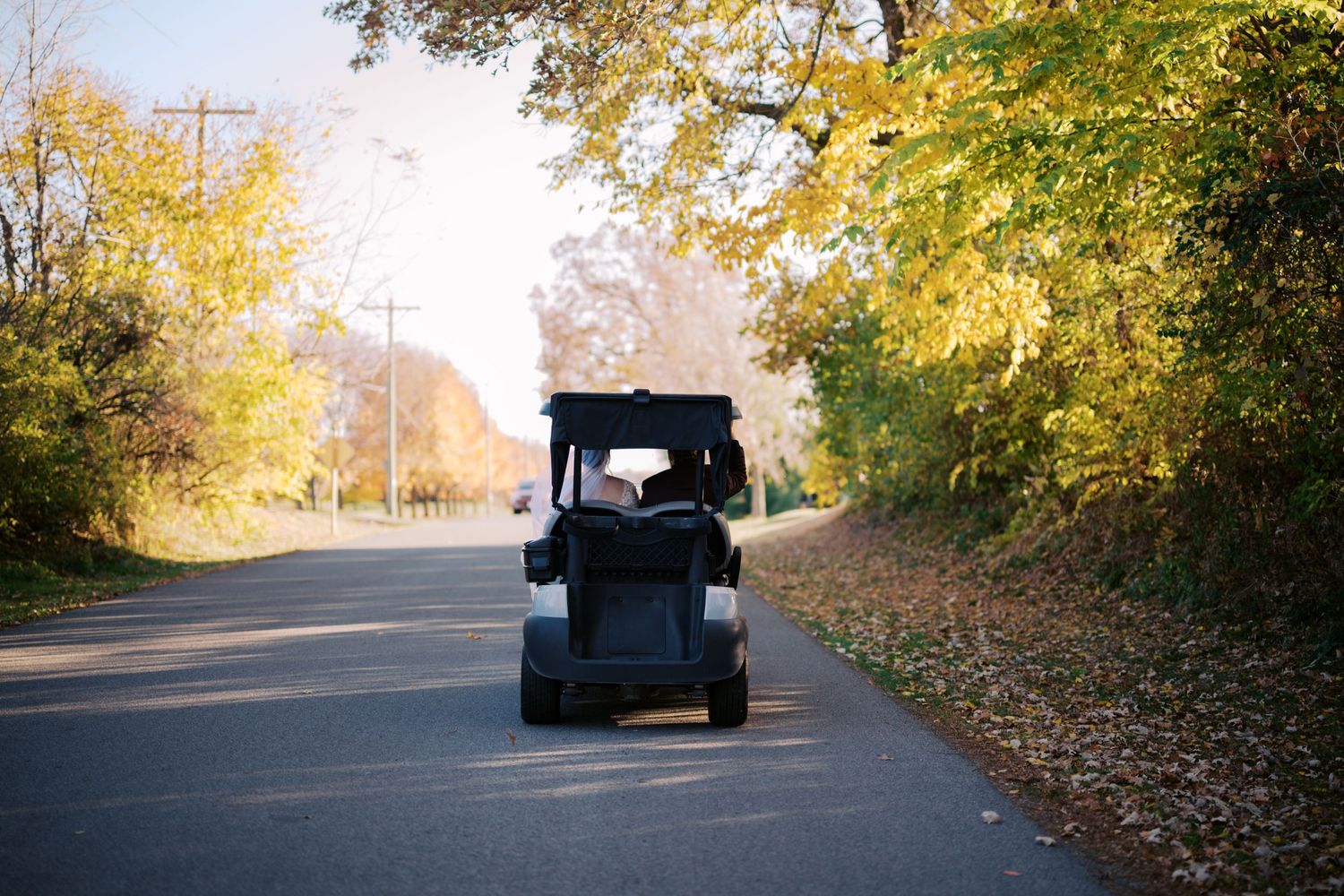 Small electric golf cart drives on a paved path surrounded by autumn trees with yellow foliage.