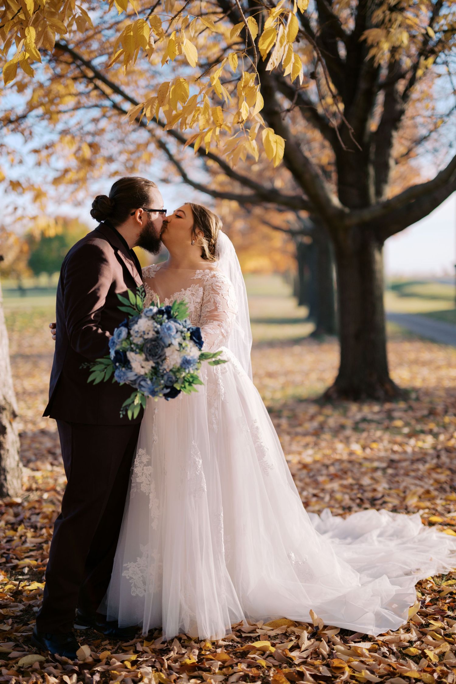 A couple sharing a romantic kiss under a golden autumn tree with fallen leaves as the bride holds a blue and purple bouquet.