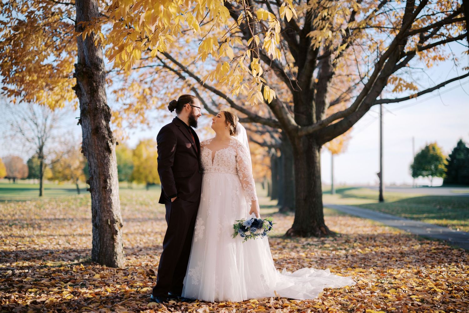 A couple in formal wedding attire share a tender moment under autumn trees with golden leaves falling around them.