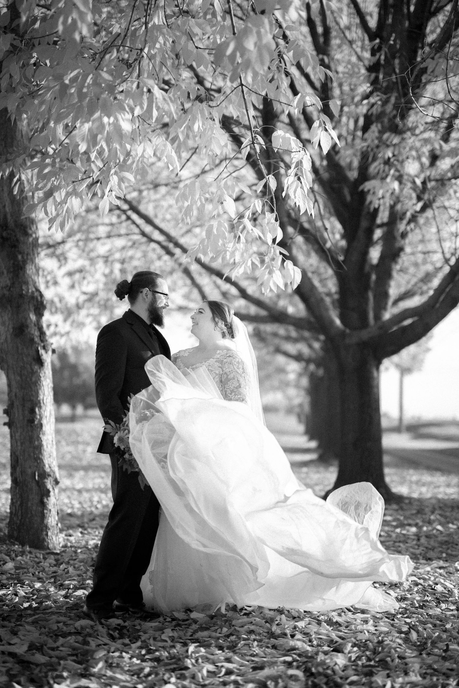 A romantic black and white wedding photo showing a couple dancing among autumn trees with the bride's dress flowing.
