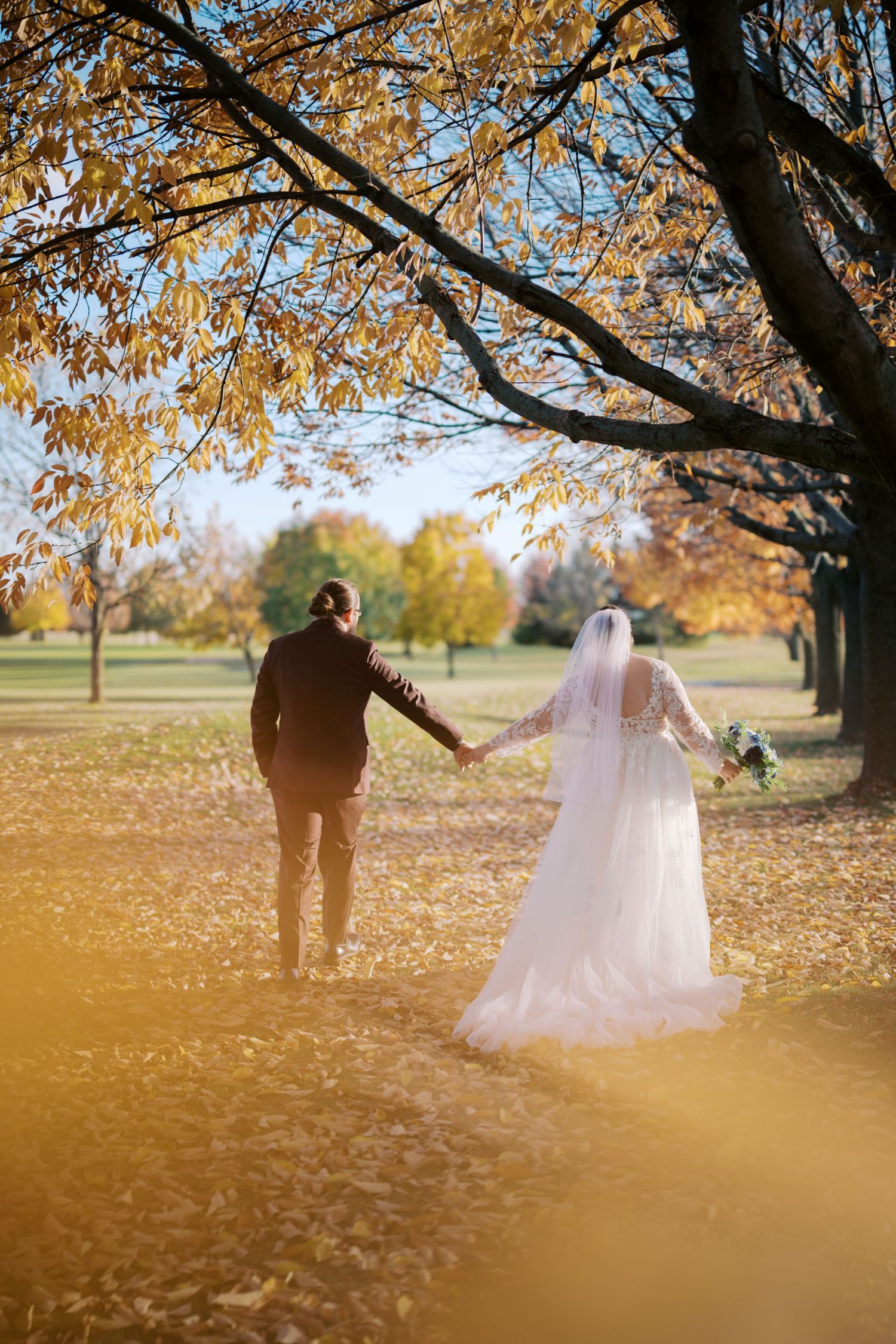 A couple in wedding attire holds hands while walking through a golden autumn pathway lined with trees.