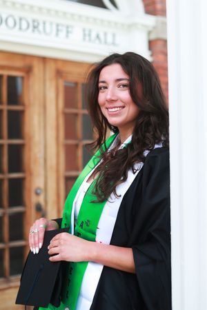 Smiling female graduate in black gown and green sash poses outside Woodruff Hall on graduation day.