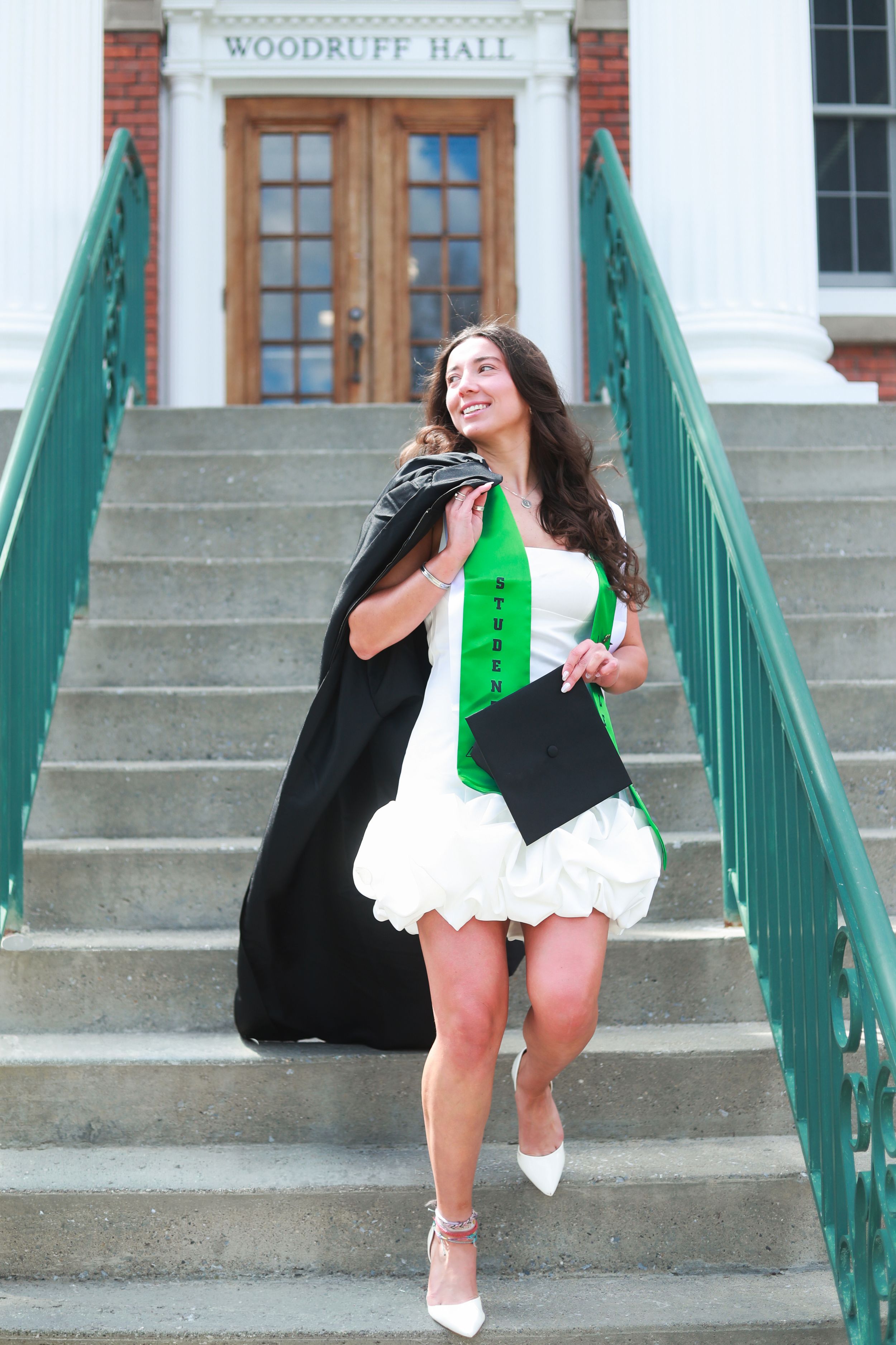 Smiling graduate in white dress with green sash holds cap and gown on Woodruff Hall steps.