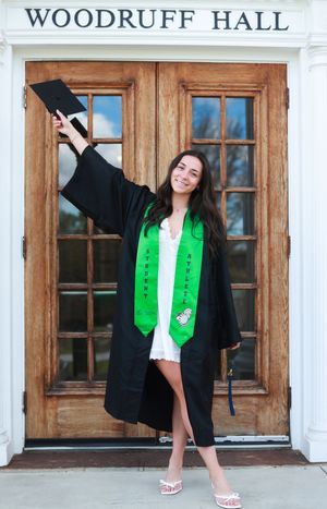 Smiling graduate in black cap and gown with green student-athlete stole posing at Woodruff Hall entrance.