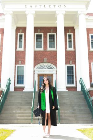 Graduate in black gown and green stole stands before Castleton University's Woodruff Hall on a sunny day.