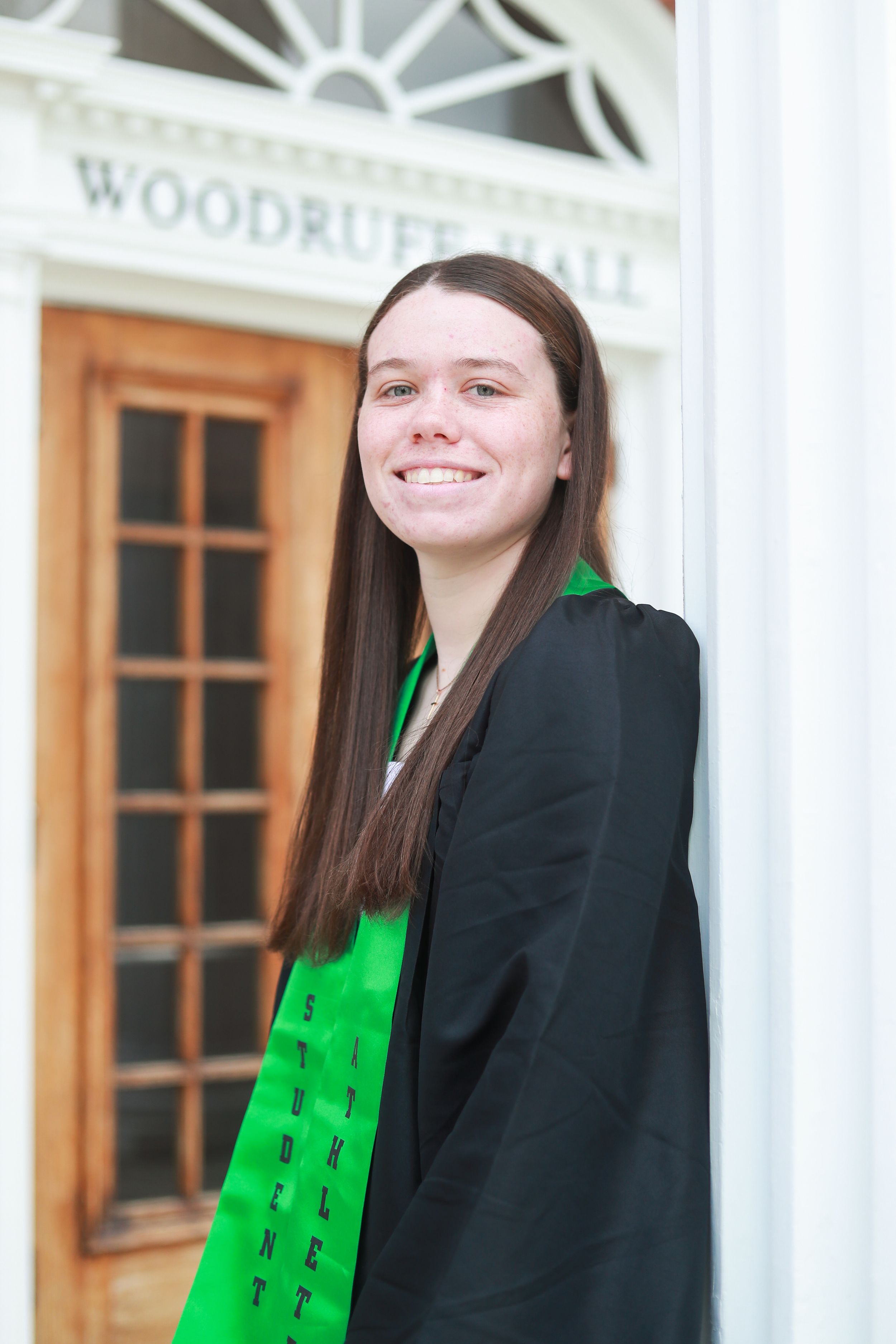 Smiling graduate in black gown and green stole poses by Woodruff Hall entrance column.