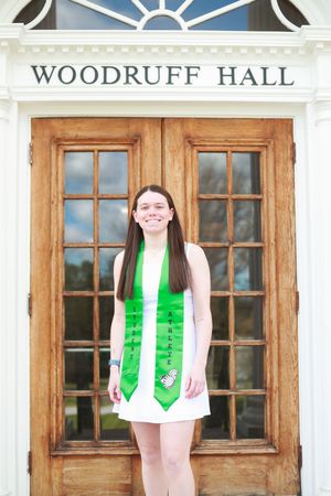Smiling graduate in white dress and green sash stands proudly before Woodruff Hall wooden doors.