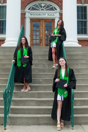 Three female graduates in black caps and gowns pose on steps outside Woodruff Hall.