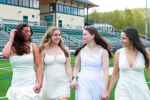 Four young women in white dresses walk hand-in-hand on a green sports field, smiling and laughing together.