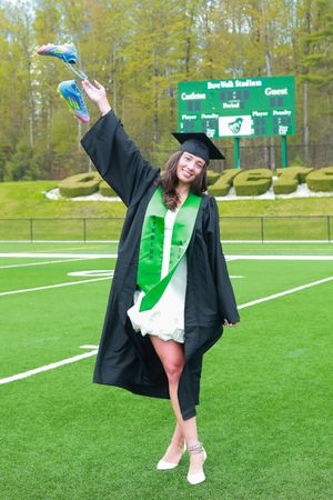 A smiling graduate in black cap and gown with green sash celebrates on a football field, holding up colorful shoes.