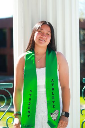 Smiling student athlete wearing a green graduation stole leaning against white columns outdoors.