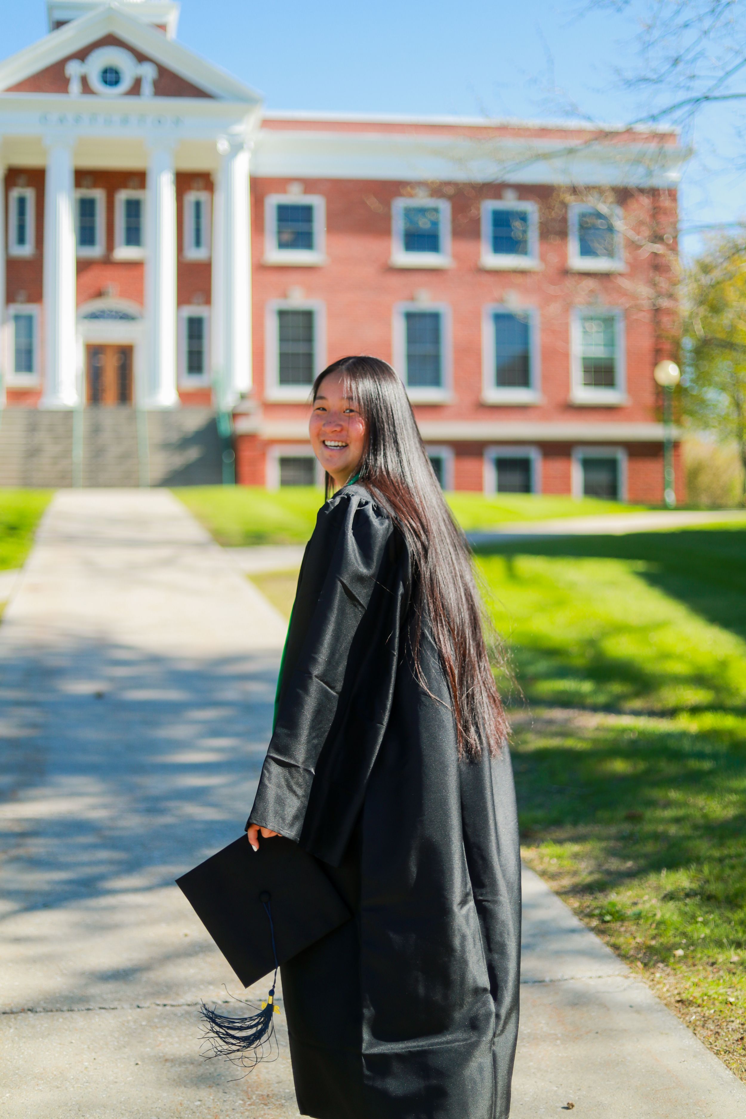 Smiling graduate in black gown holding cap on college campus path with red brick building backdrop.