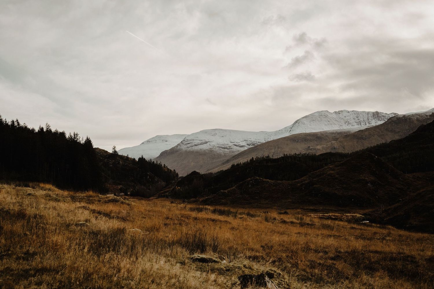 Moody mountain landscape with snow-capped peaks and golden valley in foreground.