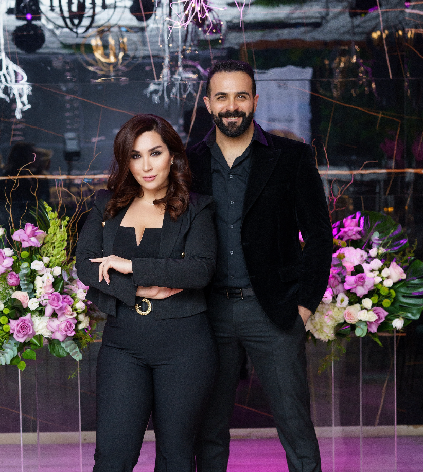 Couple in black formal attire poses with floral decorations and purple lighting.