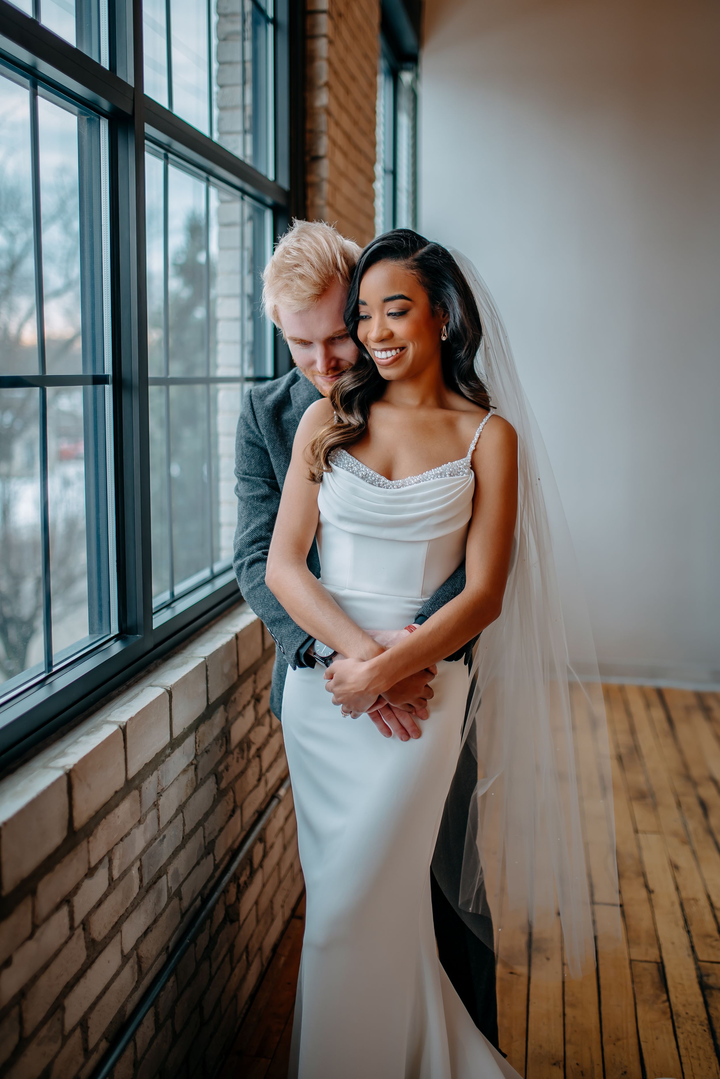 A couple shares an intimate moment by a sunlit window in a rustic brick venue on their wedding day.