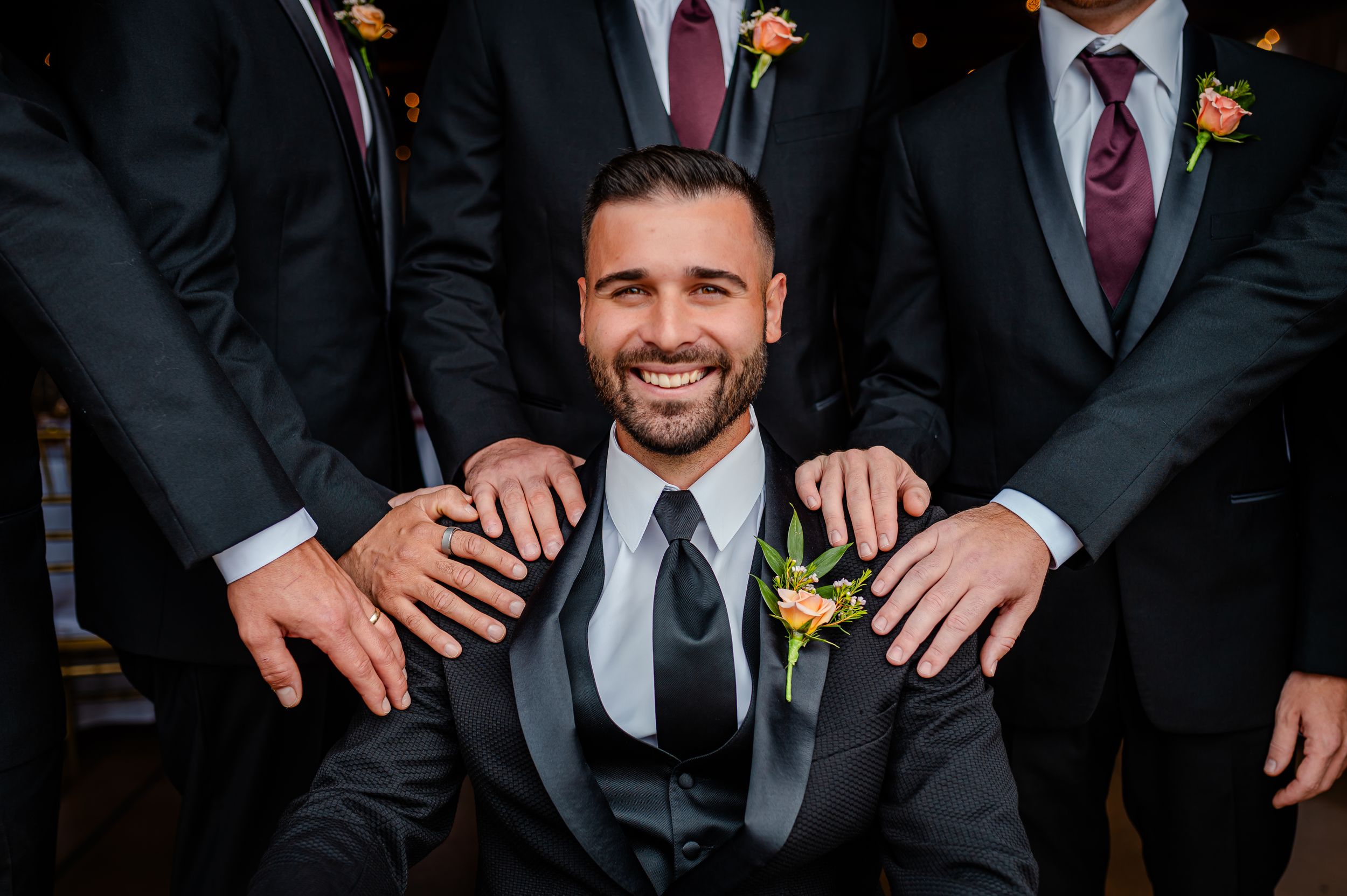 Wedding party member in black suit and tie poses while surrounded by groomsmen putting hands on his shoulders.