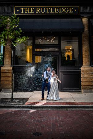 Wedding couple poses in front of The Rutledge Grand Rapids Michigan with champagne spraying on historic red brick sidewalk