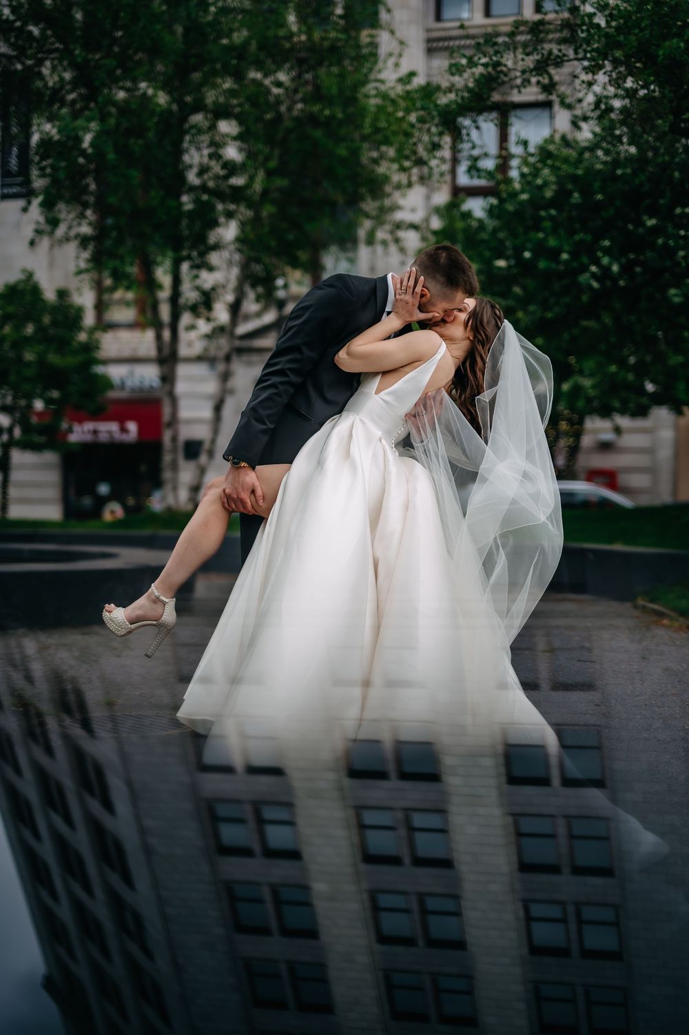 Romantic wedding couple shares tender kiss in urban setting with reflection of building and flowing white dress.