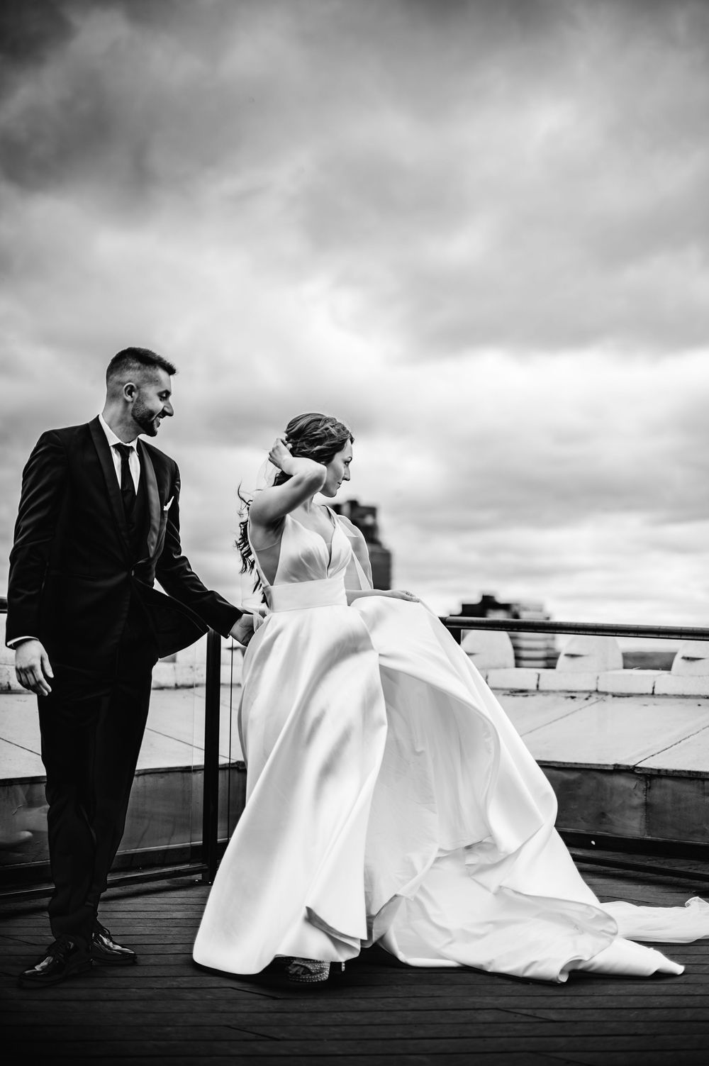A bride in a flowing white ballgown and groom in a dark suit share a moment on a rooftop terrace under dramatic clouds.
