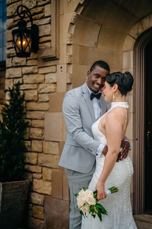 A romantic wedding couple share an intimate moment outside a stone building while holding a white floral bouquet.