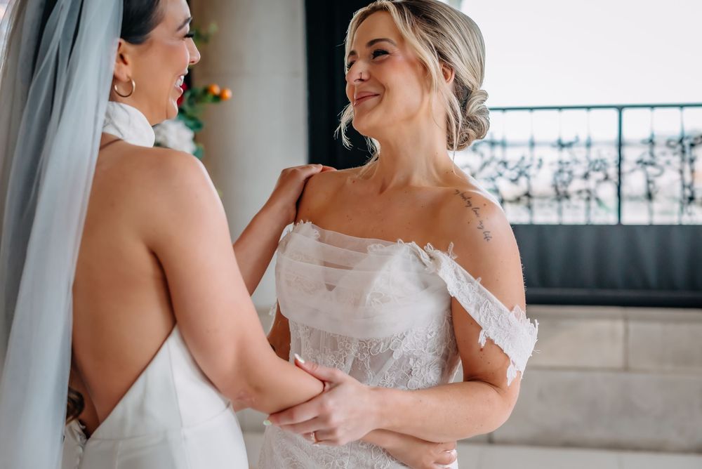 Two brides smile joyfully at each other while wearing beautiful white wedding dresses during their special day.