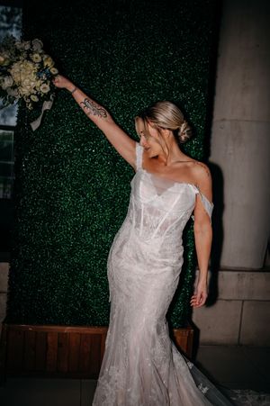 A bride wearing a white lace mermaid wedding dress raises her bouquet against a hedge wall background.