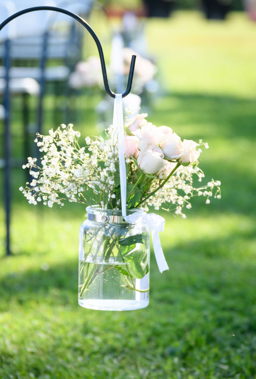 Mason jar with delicate pink roses and baby's breath hangs from shepherd's hook at outdoor wedding.