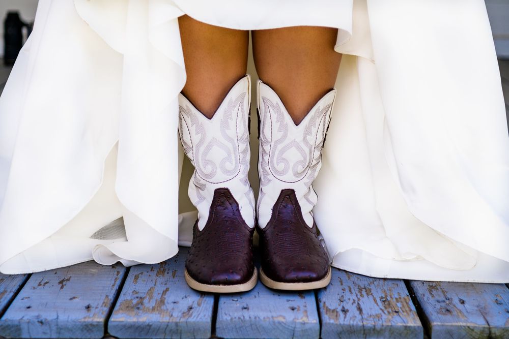 White cowboy boots peek out from under a wedding dress hem on wooden deck.