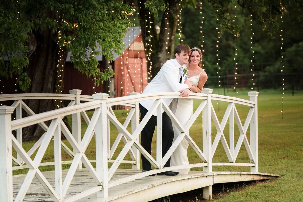 A romantic wedding couple shares an intimate moment on a white wooden bridge decorated with string lights.