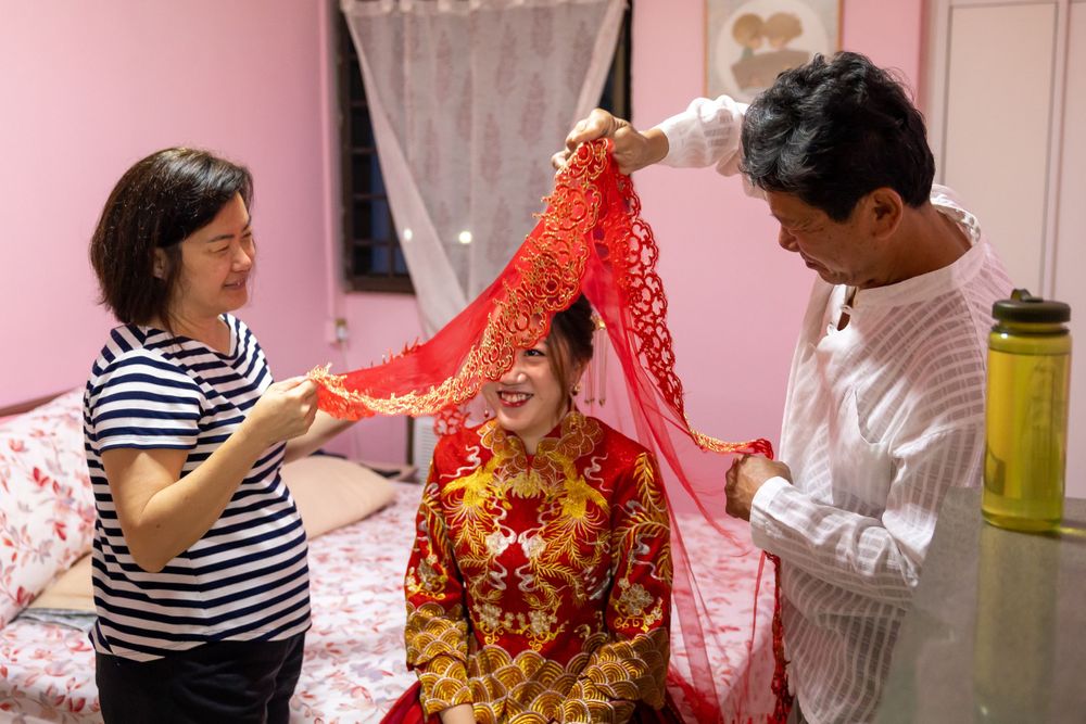 People helping adjust a red traditional Chinese wedding veil in a pink room.