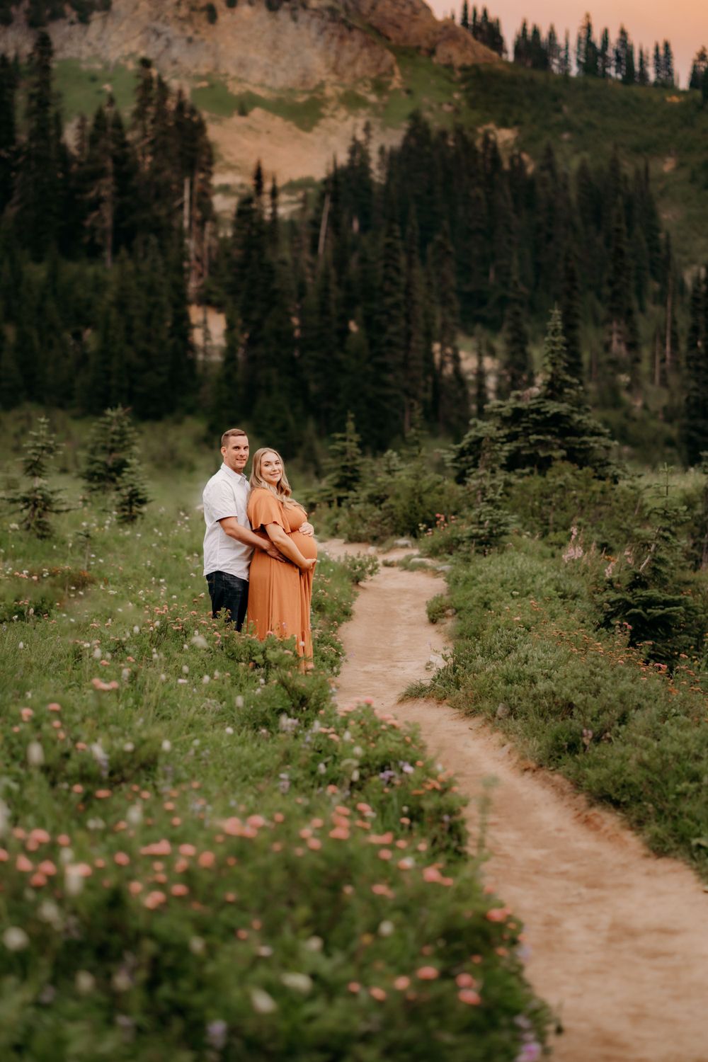 A dirt hiking trail winds through wildflowers and evergreen trees in a mountainous landscape at sunset.