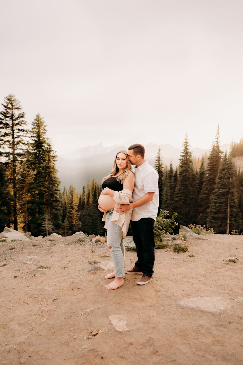 Couple embraces on mountain cliff at sunset with evergreen trees framing misty forest landscape in background for maternity photos at Tipsoo