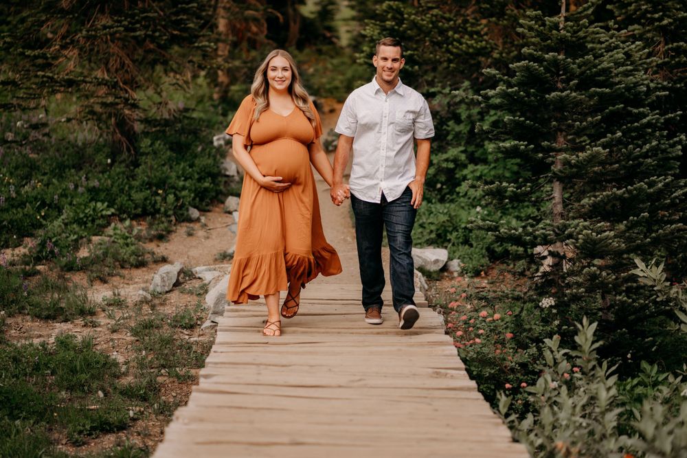 Couple walking together on wooden boardwalk path through nature for maternity photos with expectant mother wearing flowing orange dress.