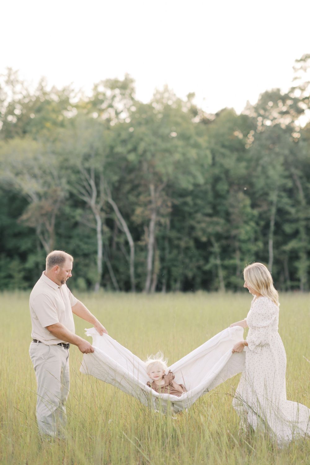 Parents swinging baby in white blanket together in a sunlit green meadow.