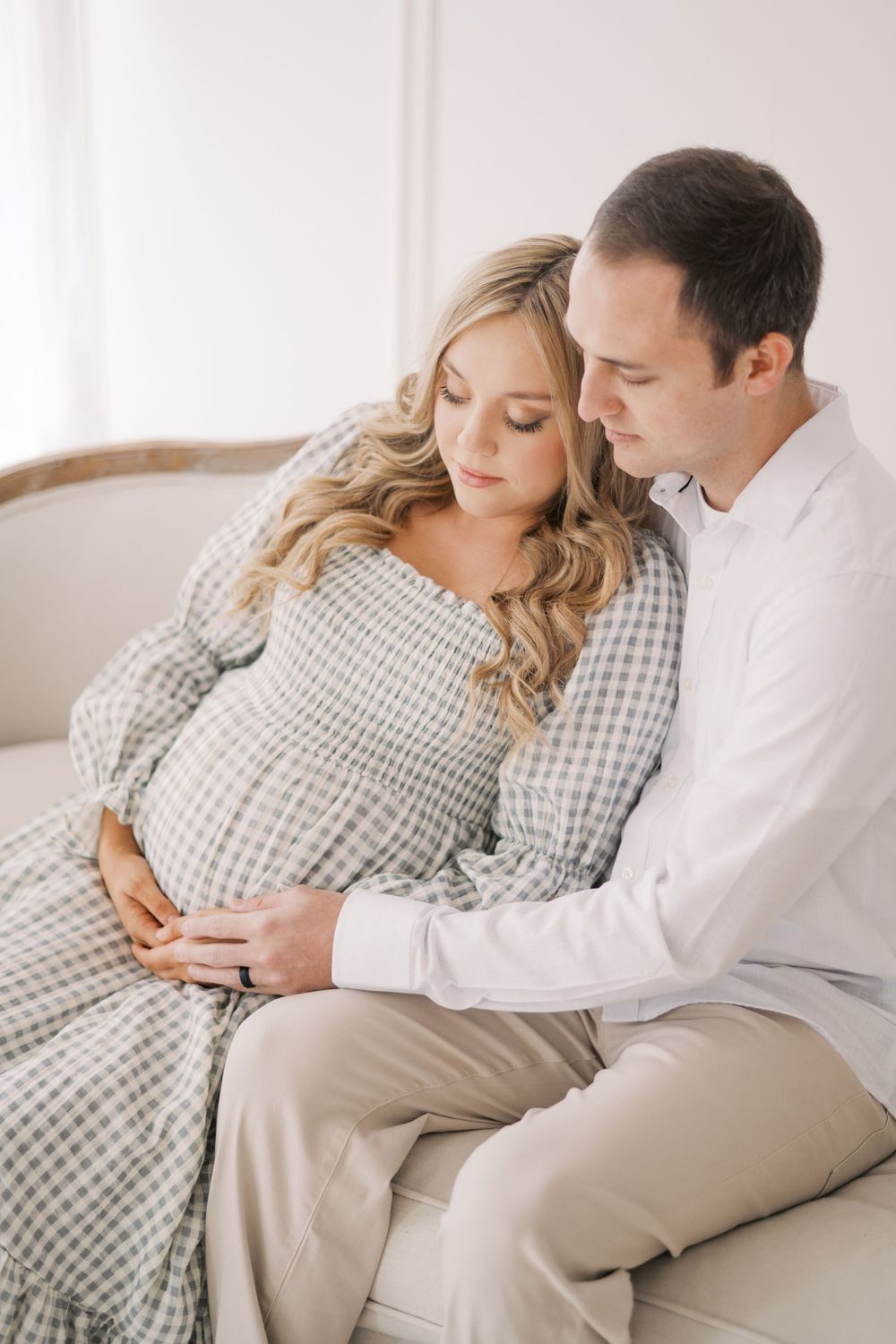 Pregnant couple embracing tenderly on a cream sofa in soft natural light.