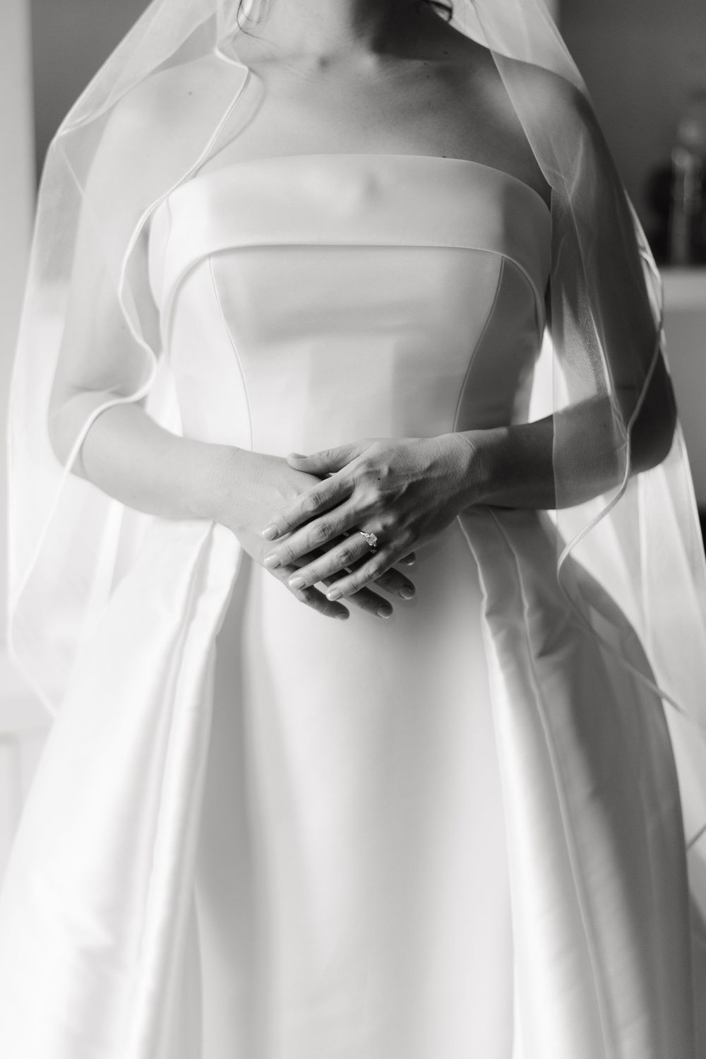 Black and white detail of an elegant wedding dress with strapless bodice and flowing skirt, showing clasped hands.