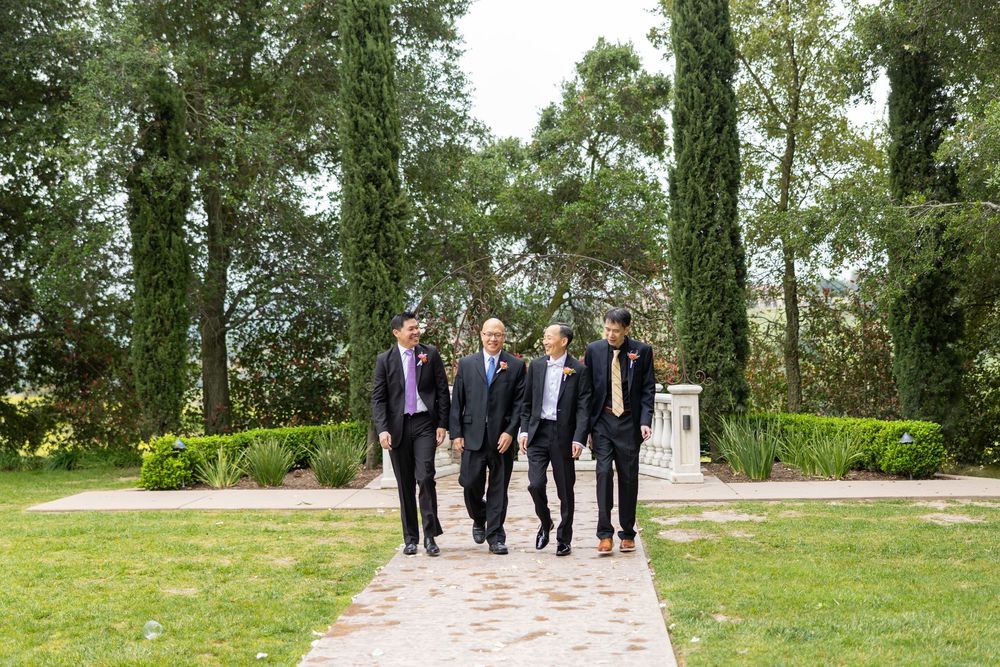 Four people walking together along a stone path in a scenic garden setting with tall trees.