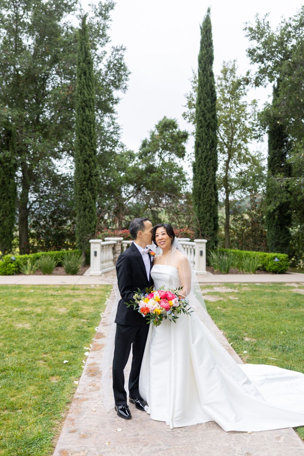 A wedding couple stands together on a garden path lined with cypress trees and lush greenery.