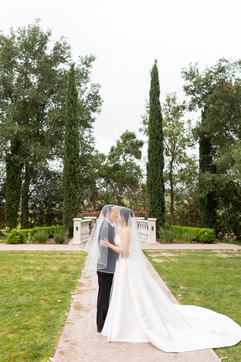 A couple embraces on a garden path surrounded by cypress trees and lush landscaping at an elegant outdoor venue.