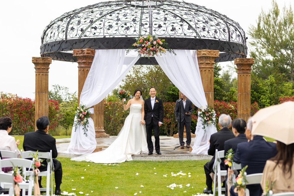 Outdoor wedding ceremony under an elegant gazebo with white draping and floral decorations, surrounded by guests on the lawn.