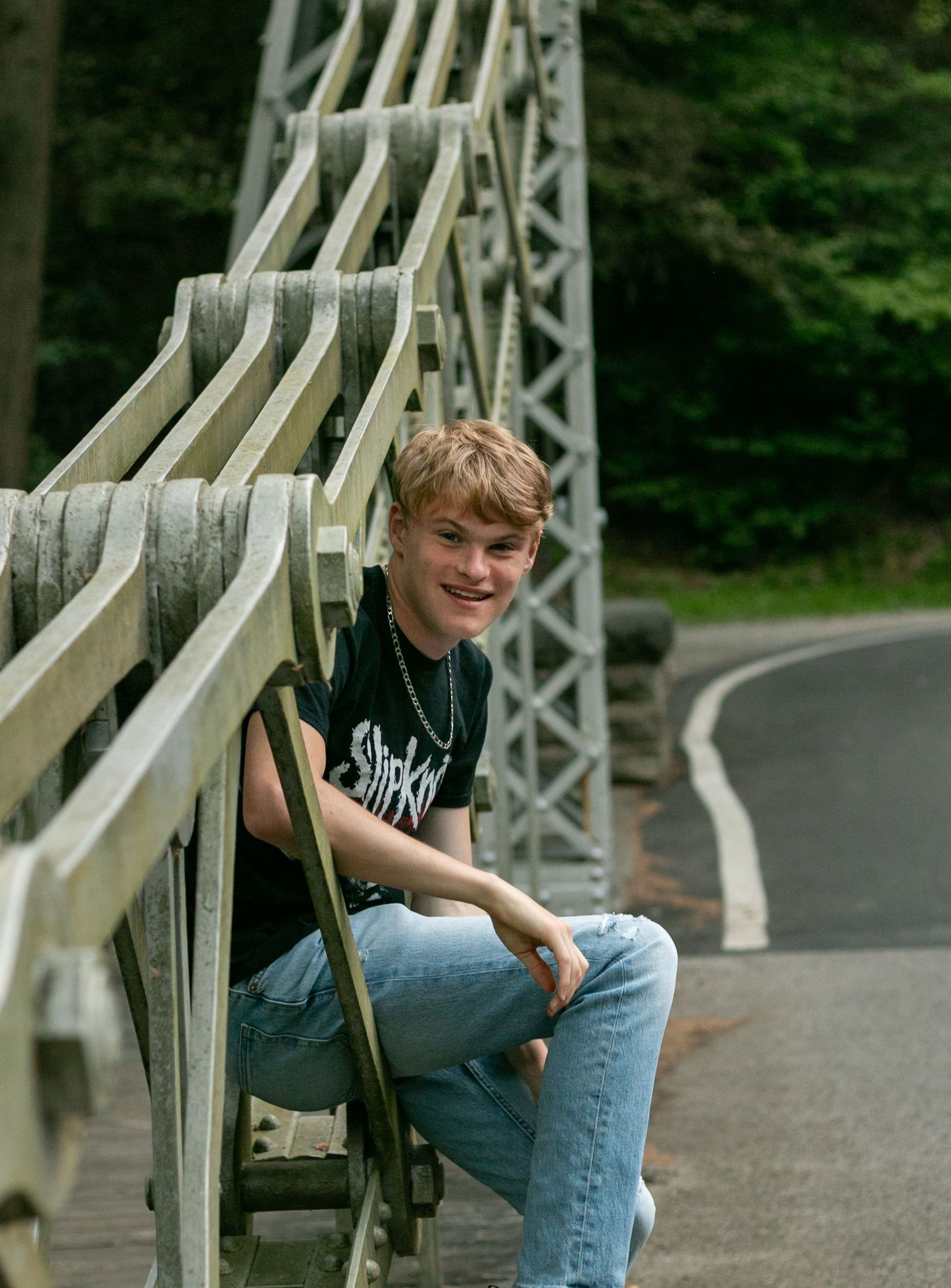A person sits casually on a white metal bridge railing along a curved road surrounded by green foliage.