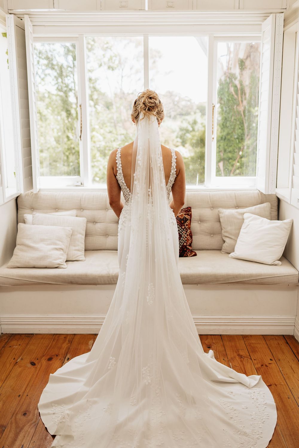 Wedding dress with delicate lace details and flowing train displayed against white window bench seating.