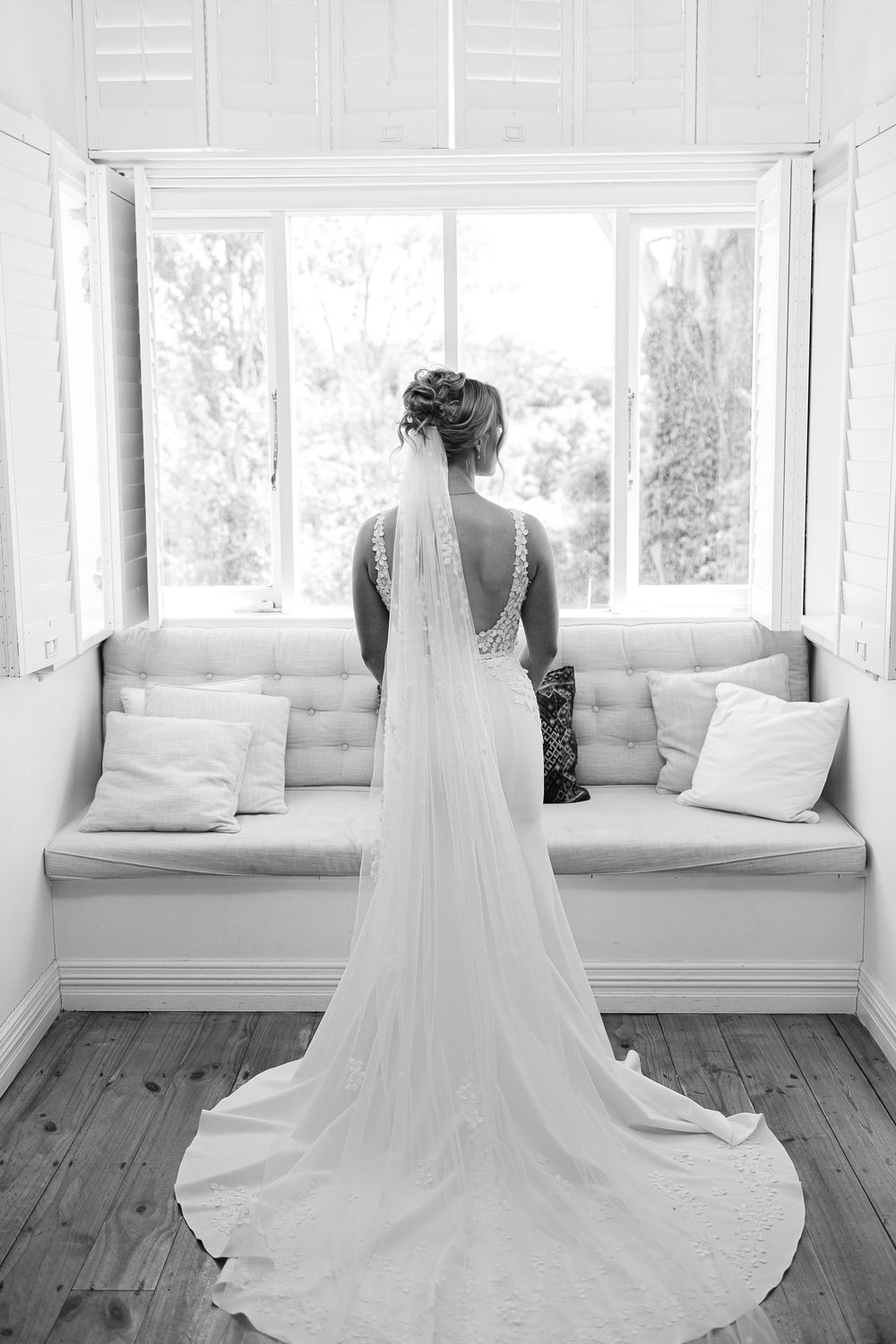 A bride in an elegant white gown with a flowing veil stands by a window seat in a sun-lit room.