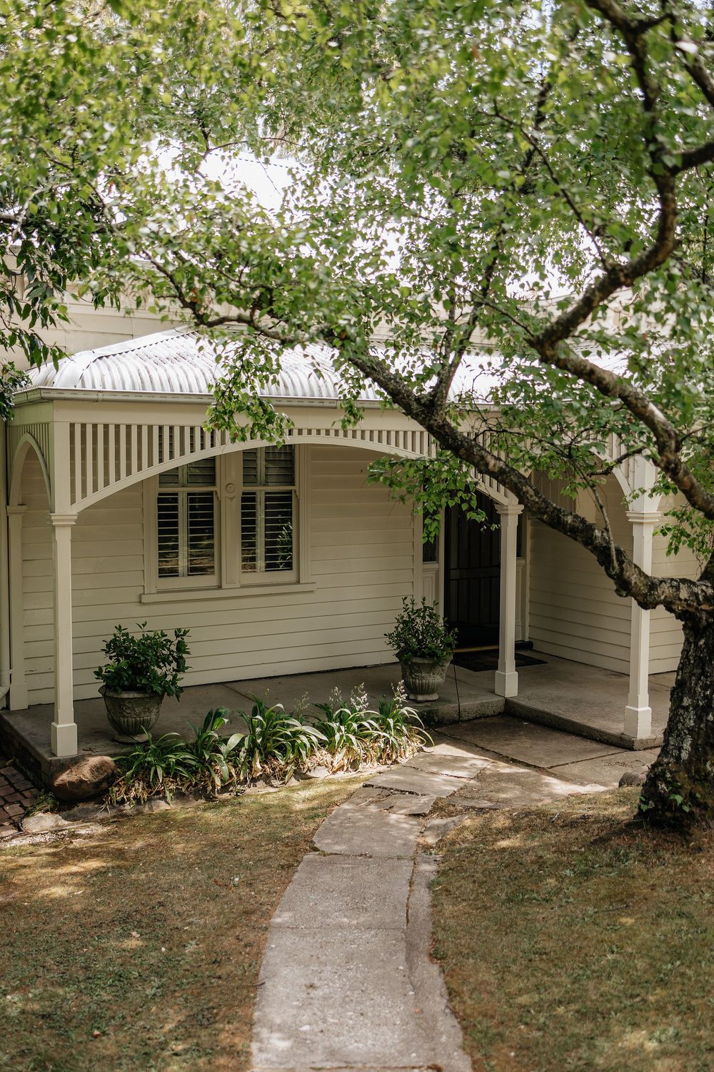 A white cottage-style house with a decorative verandah and curved concrete path leading to the entrance framed by overhanging trees.