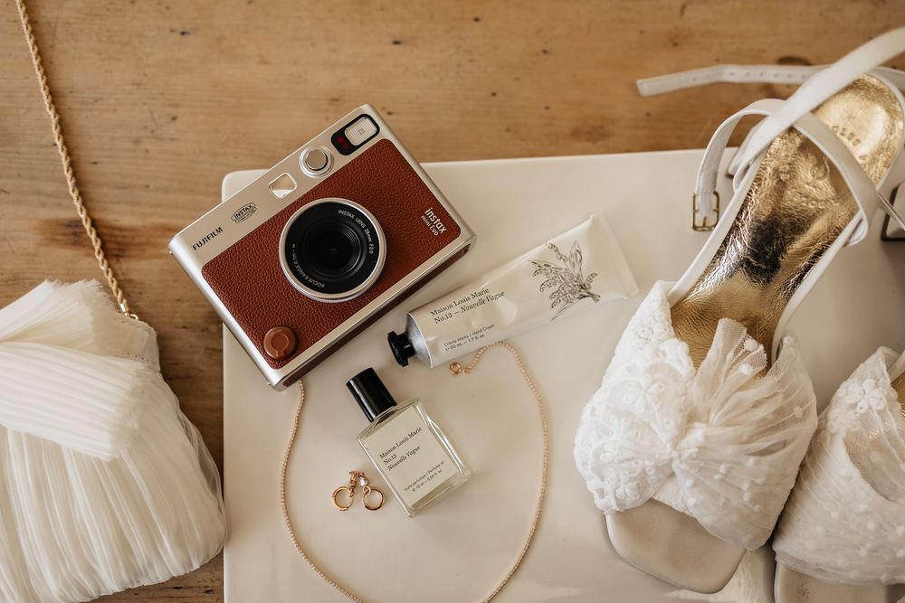 Wedding accessories flatlay with vintage camera, bridal shoes, rings and perfume bottle on wooden surface.