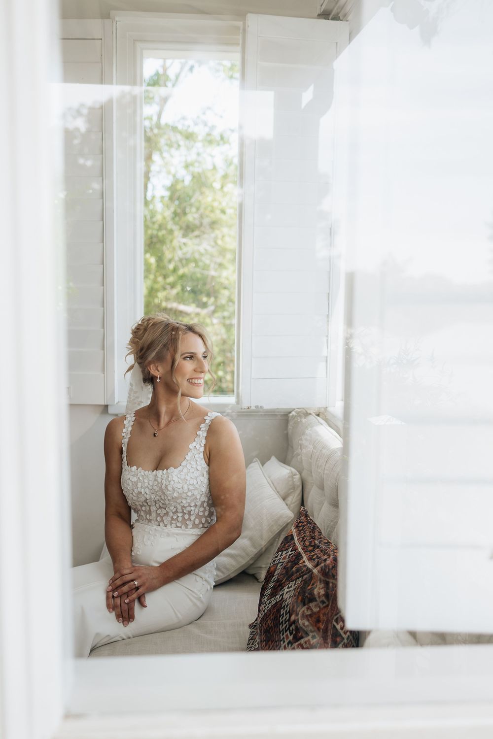 A bride in a beaded gown sits by a bright window on a white cushioned bench.