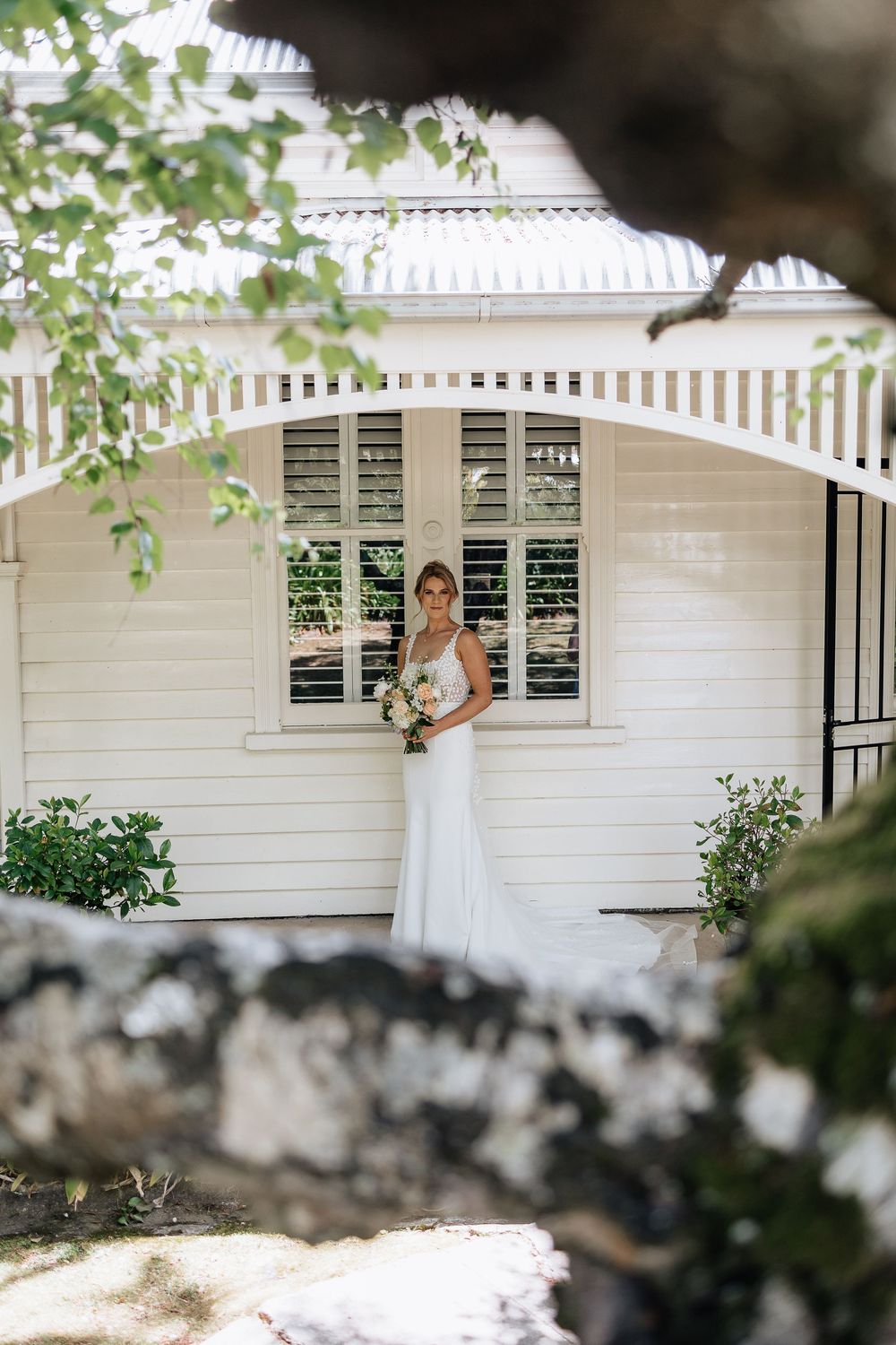 A bride in white dress holds bouquet in front of classic white cottage with shuttered windows.