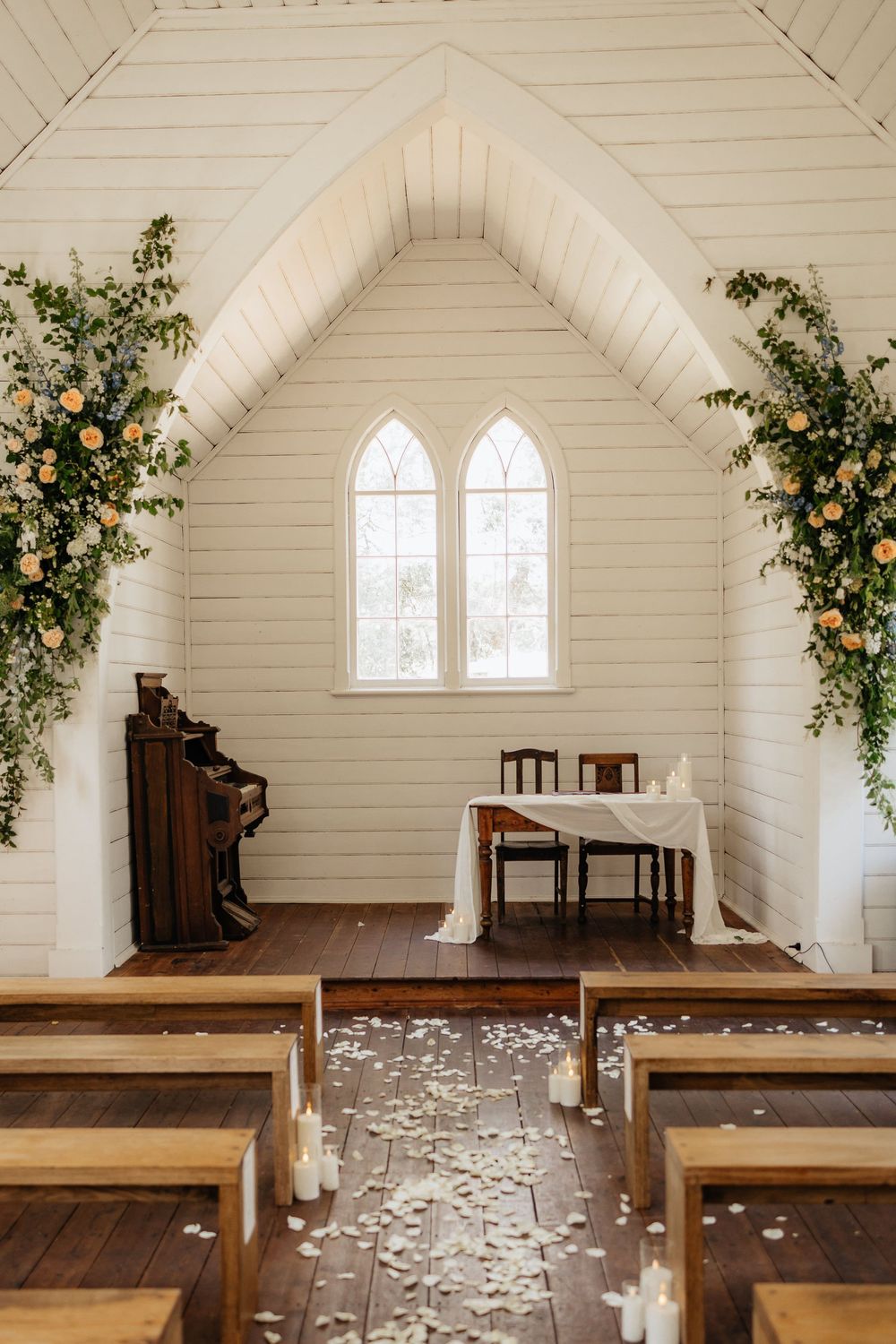 White chapel interior decorated with floral arrangements and wooden pews with scattered rose petals.