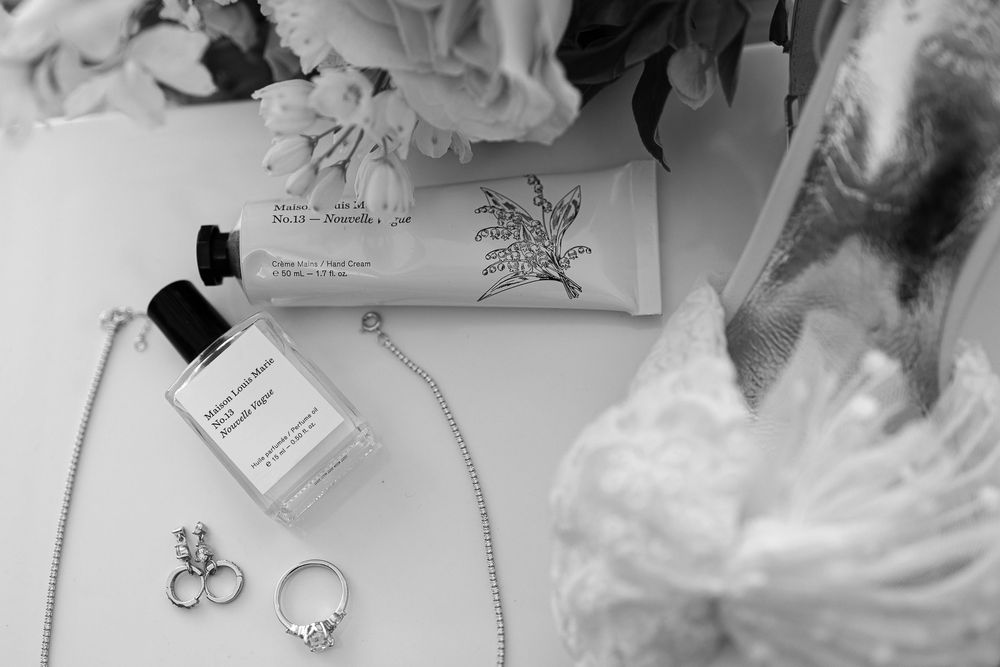 Black and white photo of wedding details including rings, perfume bottle, and delicate lace.